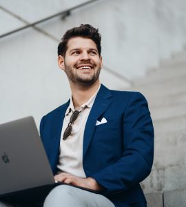 Ein lächelnder Mann in einem blauen Blazer sitzt auf einer Treppe im Freien, hat einen Laptop auf dem Schoß und blickt nach oben. Die Sonnenbrille hängt von seinem Hemd, und er wirkt entspannt und glücklich.