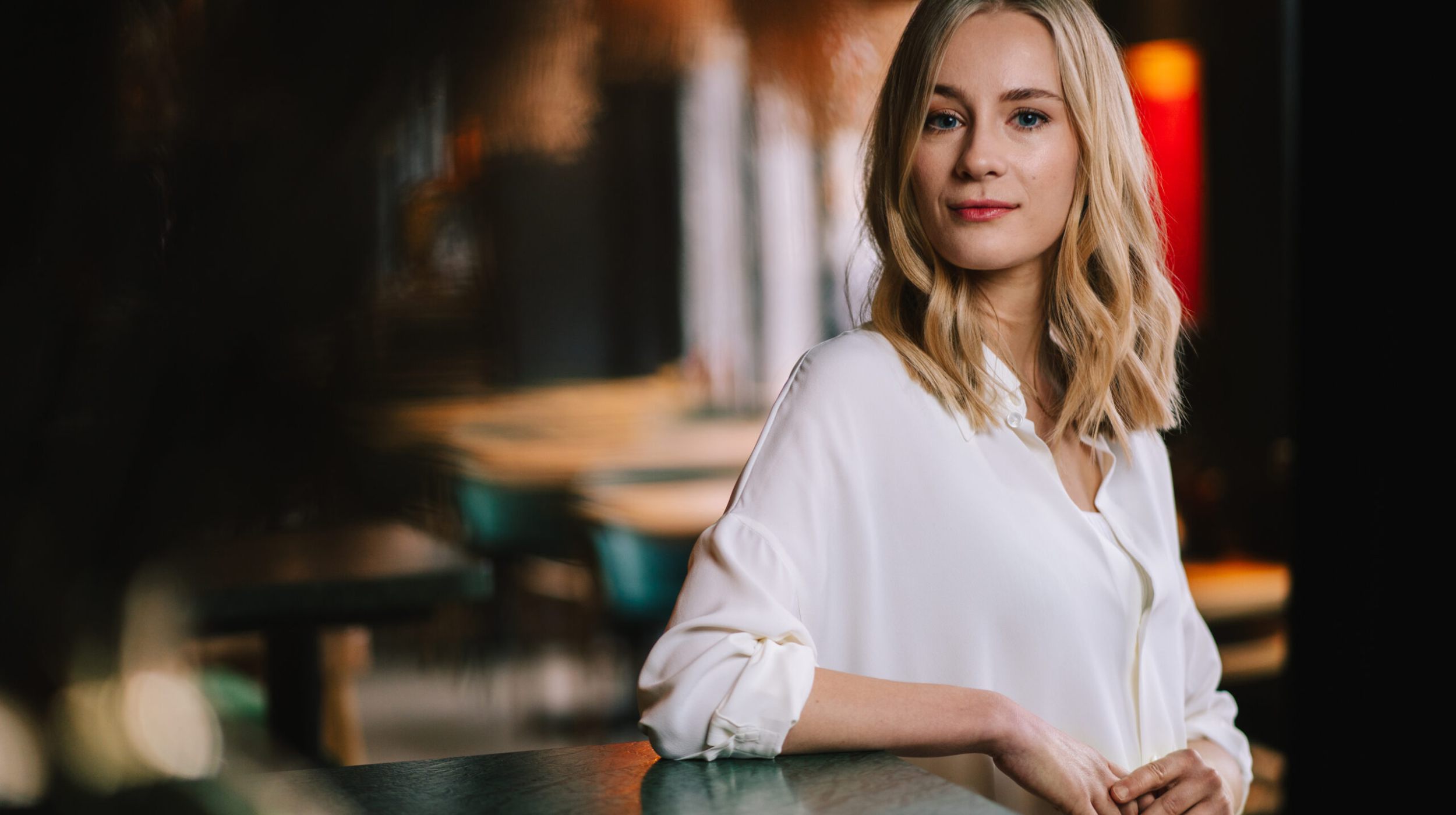 Eine Frau mit blondem Haar und weißer Bluse steht an einem grünen Tisch in einem schwach beleuchteten Innenraum und schaut selbstbewusst in die Kamera. Der Hintergrund ist unscharf, Stühle und warmes Licht sind zu sehen.