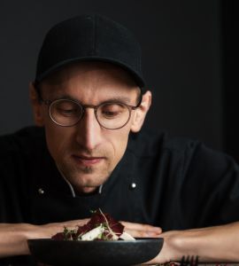 Ein Koch mit Brille und schwarzer Mütze prüft vor einem dunklen Hintergrund ein Gericht, das vor ihm steht, während er die Hände auf den Tisch stützt.