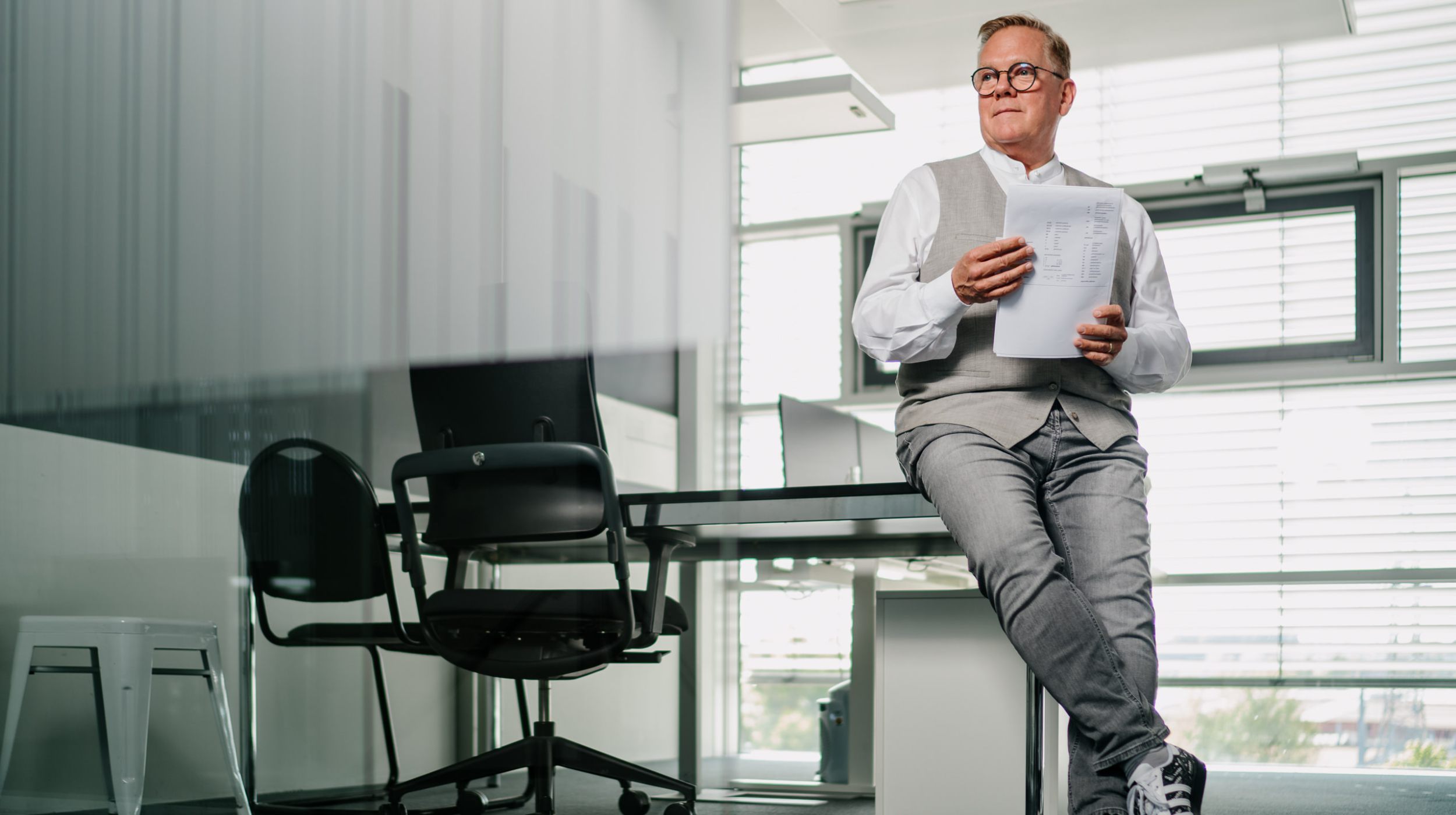 Ein Mann in legerer Geschäftskleidung sitzt am Rande eines Schreibtisches in einem modernen Büro, hält Dokumente in der Hand und schaut nachdenklich. Das Büro hat große Fenster, einen Stuhl und ist durch natürliches Licht gut beleuchtet.