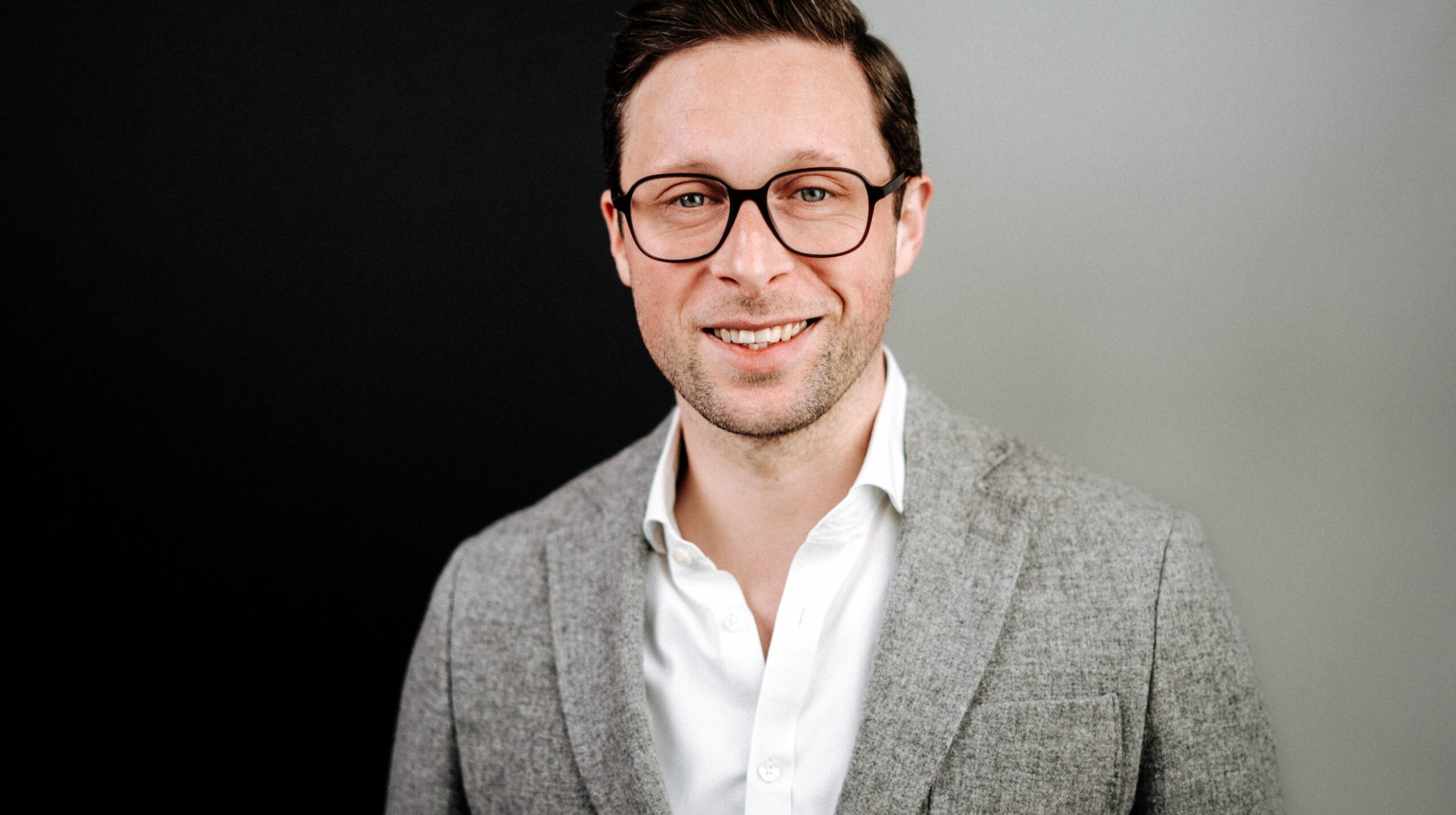 Ein Mann, der eine Brille, einen grauen Blazer und ein weißes Hemd trägt, lächelt in die Kamera. Der Hintergrund ist auf der linken Seite schwarz und auf der rechten Seite hellgrau.