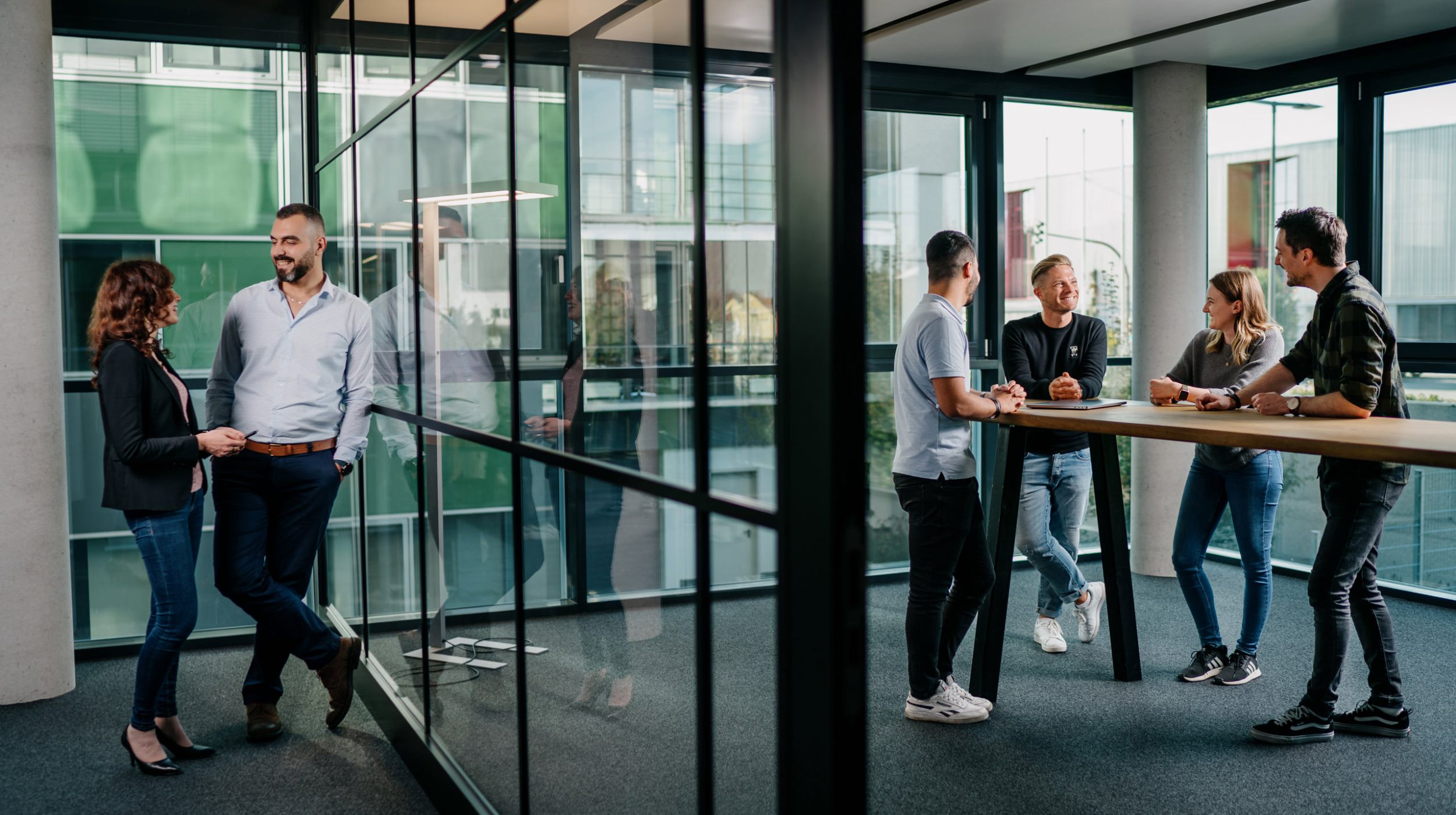 Zwei Gruppen von Personen in legerer Geschäftskleidung unterhalten sich in einem modernen Büro mit großen Fenstern und Glaswänden; ein Paar steht in der Nähe des Fensters, und vier andere unterhalten sich an einem hohen Tisch.