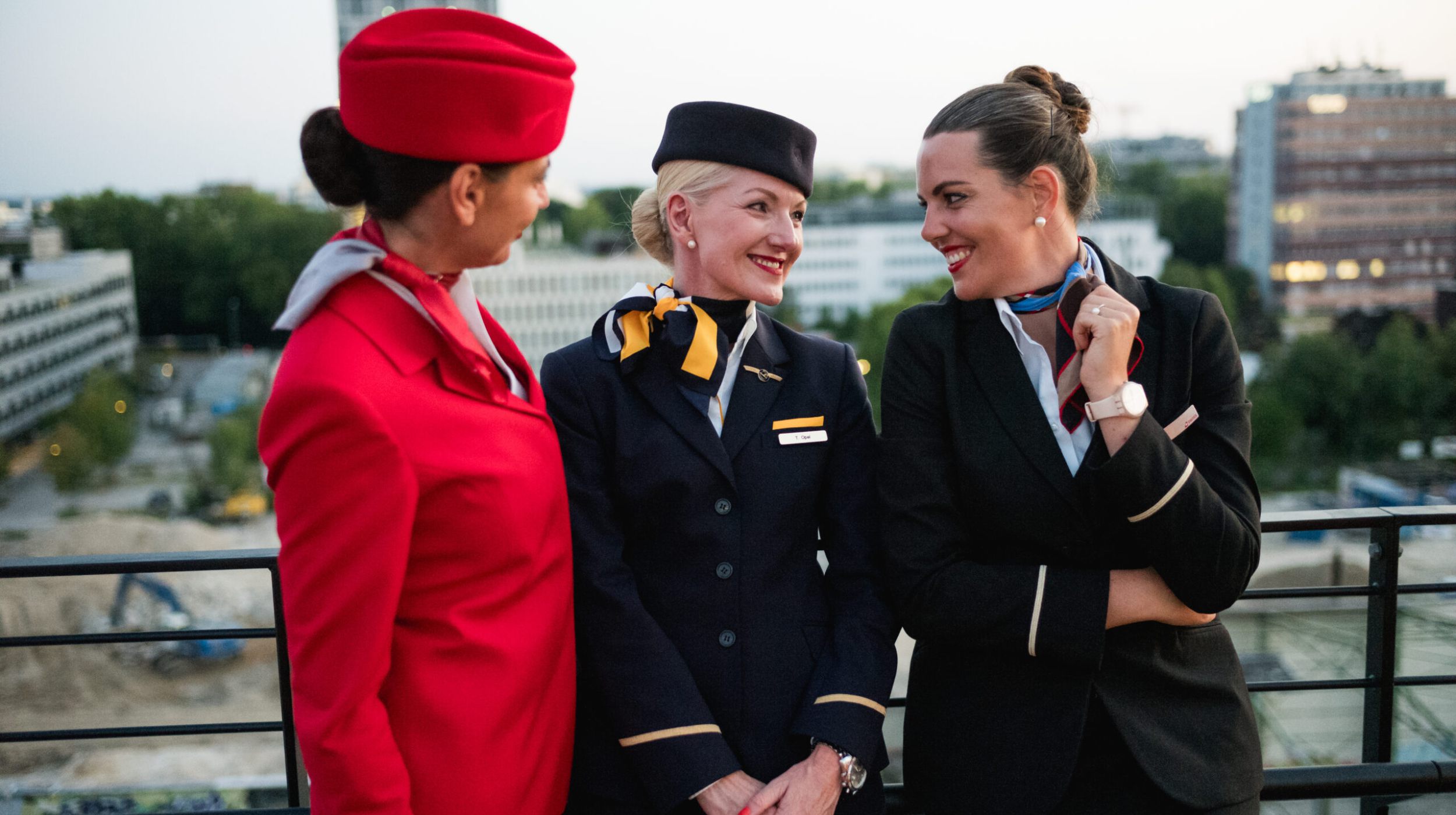 Drei Frauen in verschiedenen Uniformen von Fluggesellschaften stehen zusammen auf einem Dach, lächeln und unterhalten sich. Im Hintergrund sind städtische Gebäude und Grünanlagen zu sehen.