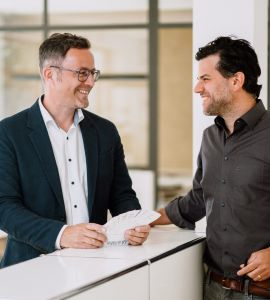 Zwei Männer stehen und lächeln, während sie sich in einem Büro unterhalten; einer hält einen Stapel Papiere, und beide scheinen ein freundliches Gespräch zu führen.
