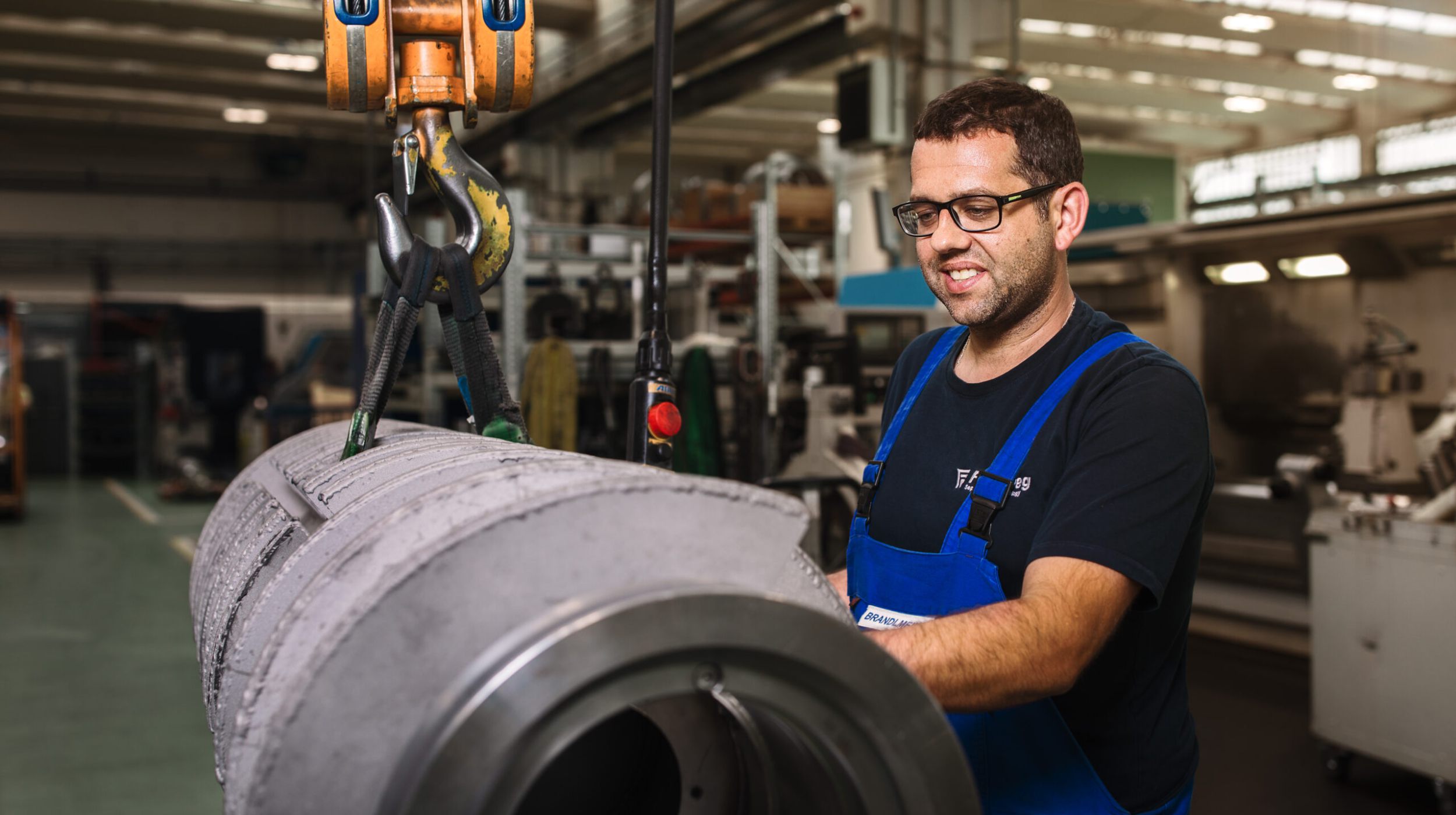 Ein Mann mit Brille und blauem Overall bedient einen Kran, um ein großes zylindrisches Maschinenteil in einer Industriehalle anzuheben. Er lächelt und konzentriert sich auf seine Aufgabe.