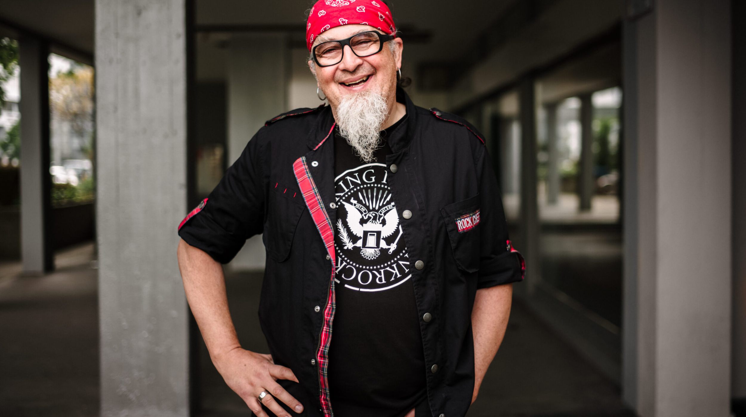 Ein lächelnder Mann mit weißem Spitzbart trägt eine Brille, ein rotes Halstuch und ein schwarzes T-Shirt mit einer Grafik unter einer offenen schwarzen Jacke. Er steht im Freien, die Hand in die Hüfte gestemmt, vor einer Betonsäule und einem Gebäudesteg.
