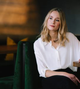 Eine Frau mit blondem Haar und weißer Bluse sitzt an einem Holztisch in einem schwach beleuchteten Restaurant und schaut nachdenklich in die Ferne.