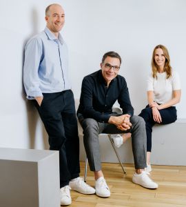 Drei Personen posieren in einem minimalistischen Raum. Zwei Männer, einer stehend und einer sitzend, und eine Frau, die auf einer Bank sitzt, alle in Freizeitkleidung und weißen Turnschuhen, lächeln in die Kamera vor einer weißen Wand und einem Holzboden.