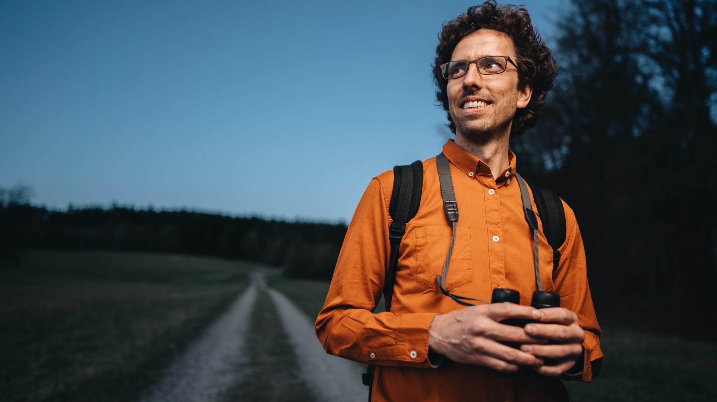 Ein Mann mit Brille und orangefarbenem Hemd steht mit einem Fernglas in der Hand auf einem Feldweg, mit einem Rucksack auf den Schultern und Bäumen im Hintergrund unter einem klaren Himmel.