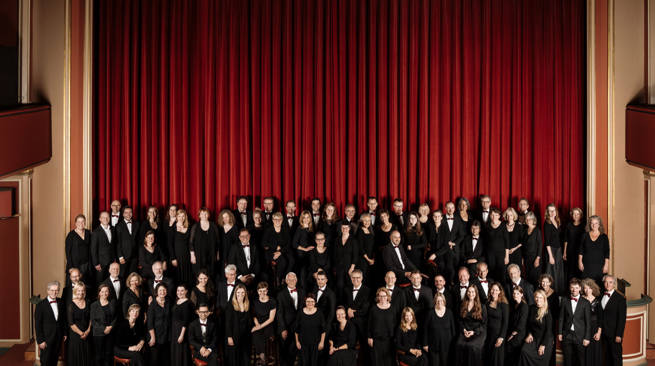 Ein großes Orchester in formeller schwarzer Kleidung steht auf der Bühne vor einem roten Vorhang und posiert für ein Gruppenfoto in einem Theater mit roten Wänden und Sitzen.