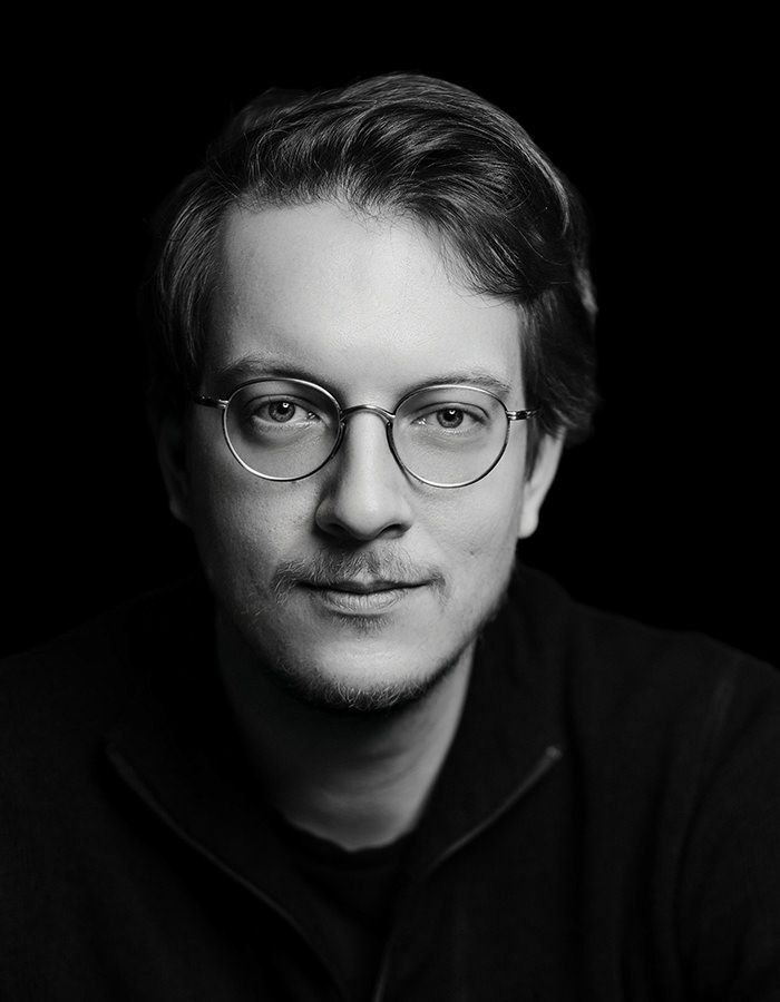 Schwarz-Weiß-Porträt einer Person mit kurzem, hellem Haar und Brille, die mit neutralem Gesichtsausdruck direkt in die Kamera schaut, vor einem dunklen Hintergrund.