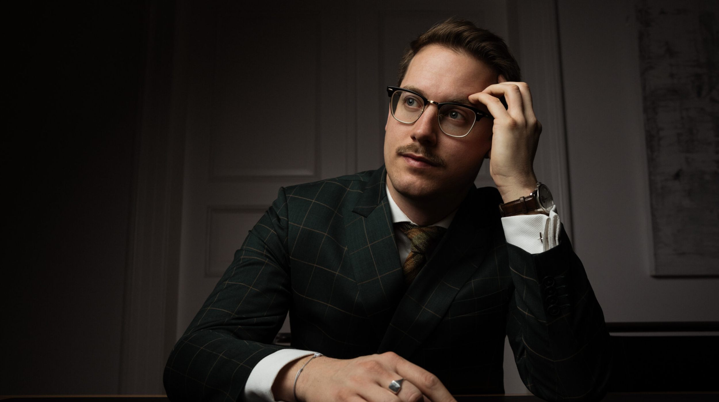 Ein Mann in einem dunkelgrün karierten Anzug und mit Brille sitzt an einem Schreibtisch, stützt seinen Kopf auf eine Hand und blickt nachdenklich nach oben. Die Beleuchtung ist gedämpft und schafft eine stimmungsvolle Atmosphäre.
