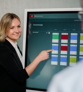 Eine Frau in Geschäftskleidung lächelt und zeigt auf einen großen Touchscreen, auf dem ein farbcodierter Zeitplan angezeigt wird, während zwei Kollegen im Vordergrund zuhören.