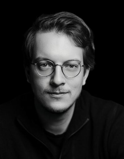 Schwarz-Weiß-Porträt einer Person mit kurzem, hellem Haar und Brille, die mit neutralem Gesichtsausdruck direkt in die Kamera schaut, vor einem dunklen Hintergrund.